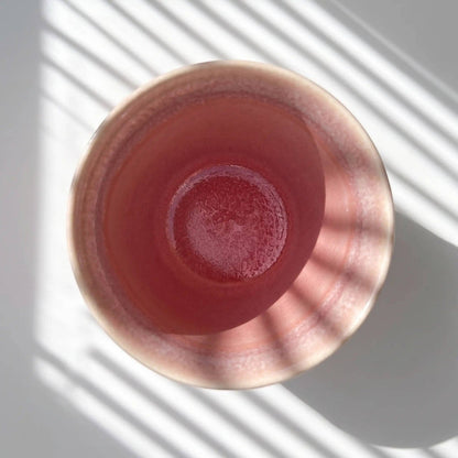 A top-down view of a pink ceramic cup or bowl with a glossy finish, set against a white background with diagonal window shadow patterns.