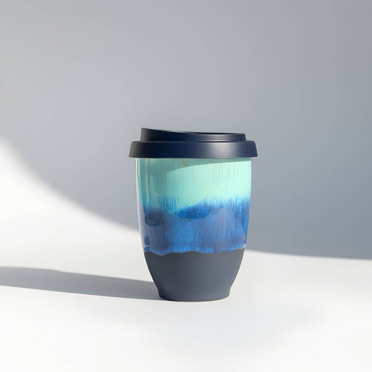 A reusable ceramic travel tumbler with a dark navy lid, featuring a gradient design with a light aqua top half and a deep blue middle section, and a solid dark navy bottom half.