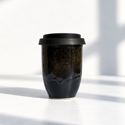 A reusable ceramic travel mug with a black lid, featuring a glossy dark brown and black glaze with a wavy lower edge, set against a bright white background with soft shadows.