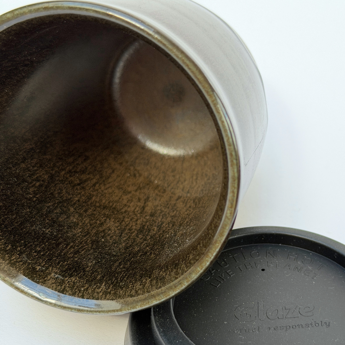 Close-up of a ceramic tumbler or mug showing the interior with a textured metallic brown and gold glaze, alongside a black lid embossed with the brand Glaze.