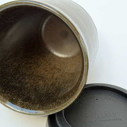 Close-up of a ceramic tumbler or mug showing the interior with a textured metallic brown and gold glaze, alongside a black lid embossed with the brand Glaze.