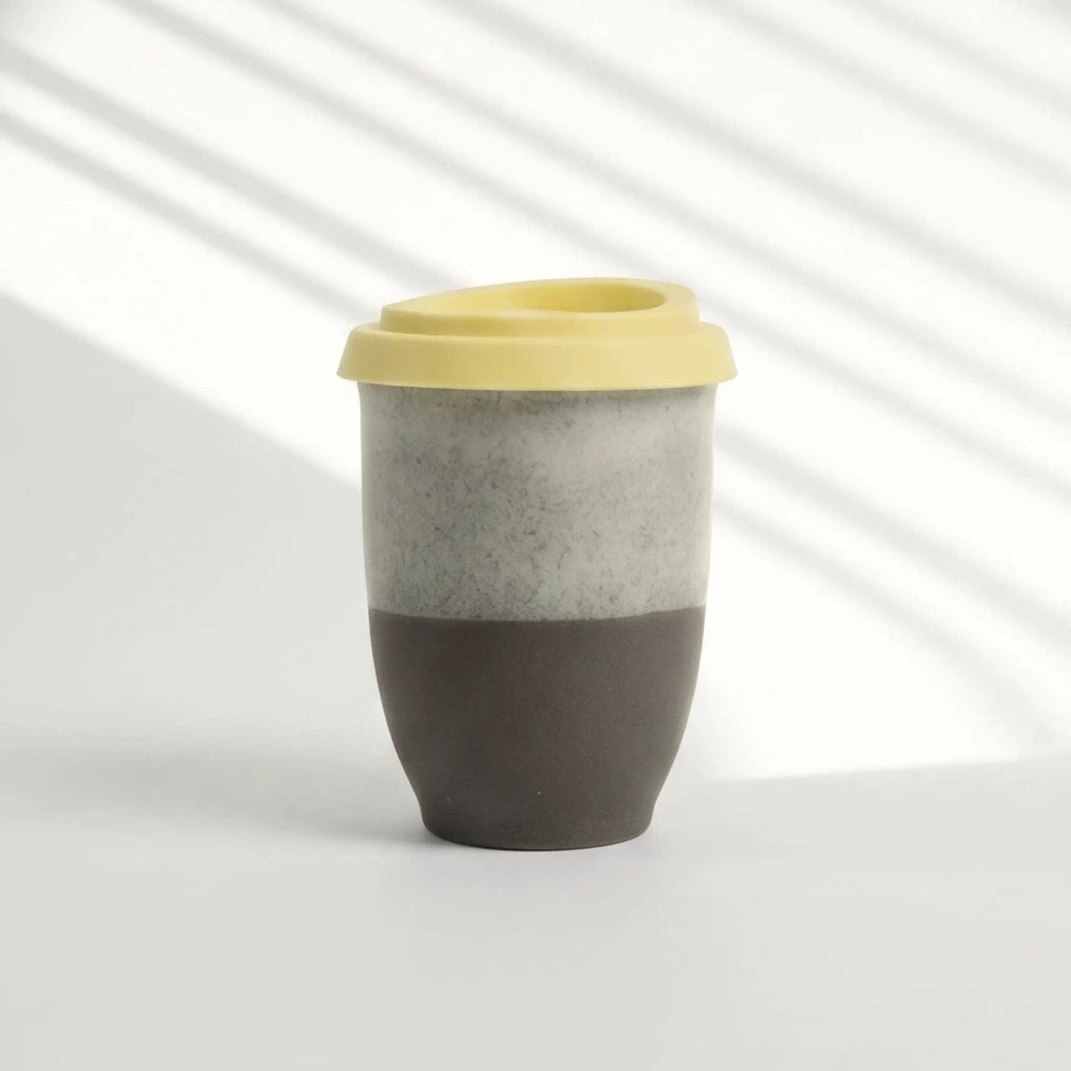A reusable travel tumbler with a yellow lid, featuring a two-tone body with a textured light gray upper half and a matte dark brown lower half, set against a white background with soft diagonal shadows.