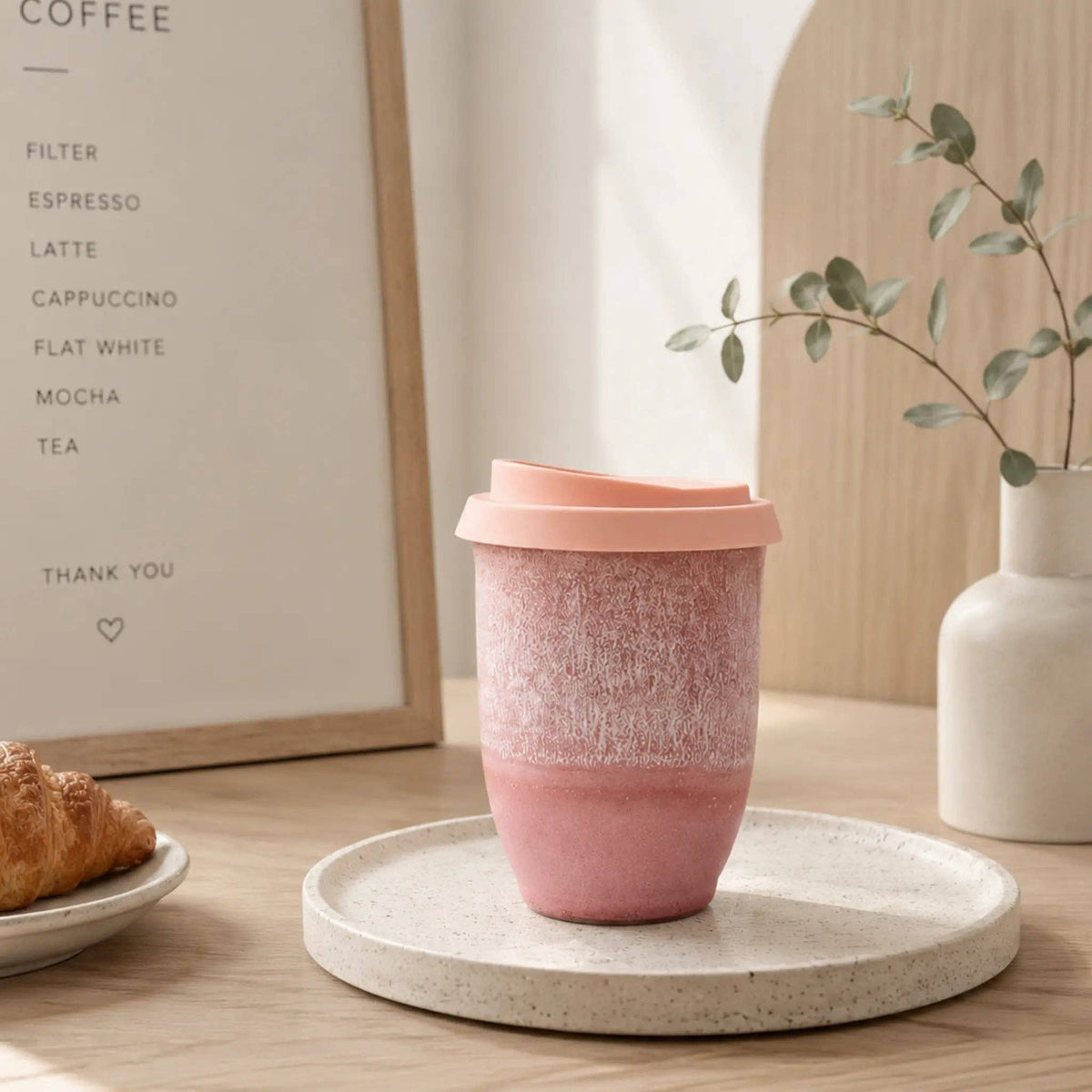 Pink ceramic cup on a white coaster with a plant and framed text in the background