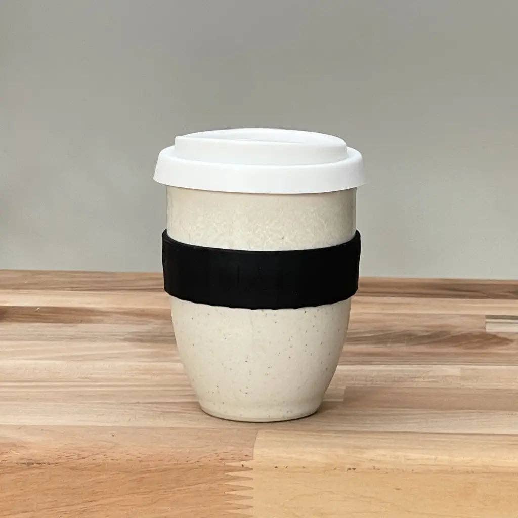 A reusable ceramic travel mug with a white lid, featuring a black fabric sleeve around the middle, set on a light wood tabletop against a neutral gray background.
