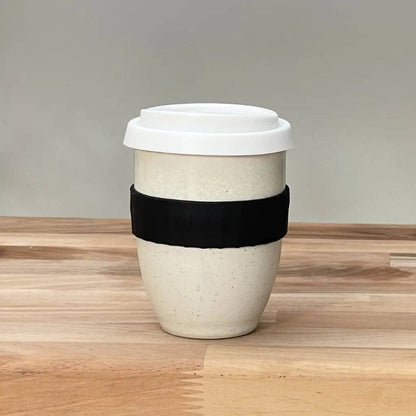 A reusable ceramic travel mug with a white lid, featuring a black fabric sleeve around the middle, set on a light wood tabletop against a neutral gray background.