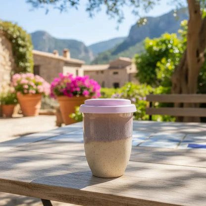 A reusable ceramic coffee cup with a light purple lid sits on a wooden table in a sunny outdoor patio setting with mountains and flowers in the background.