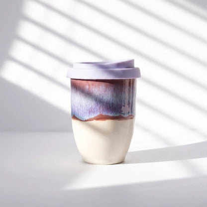 A ceramic travel mug with a lavender lid, featuring a two-tone glaze design with a cream-colored bottom half and a purple, blue, and brown marbled top half, set against a white background with diagonal window shadow patterns.