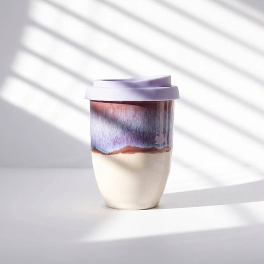 A ceramic travel mug with a lavender lid, featuring a two-tone glaze design with a cream-colored bottom half and a purple, blue, and brown marbled top half, set against a white background with diagonal window shadow patterns.