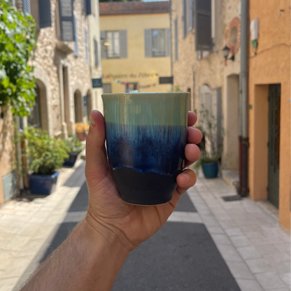 A hand holds a ceramic cup with a gradient glaze from light green to deep blue and black, set against a blurred background of a sunny stone street with buildings and potted plants.
