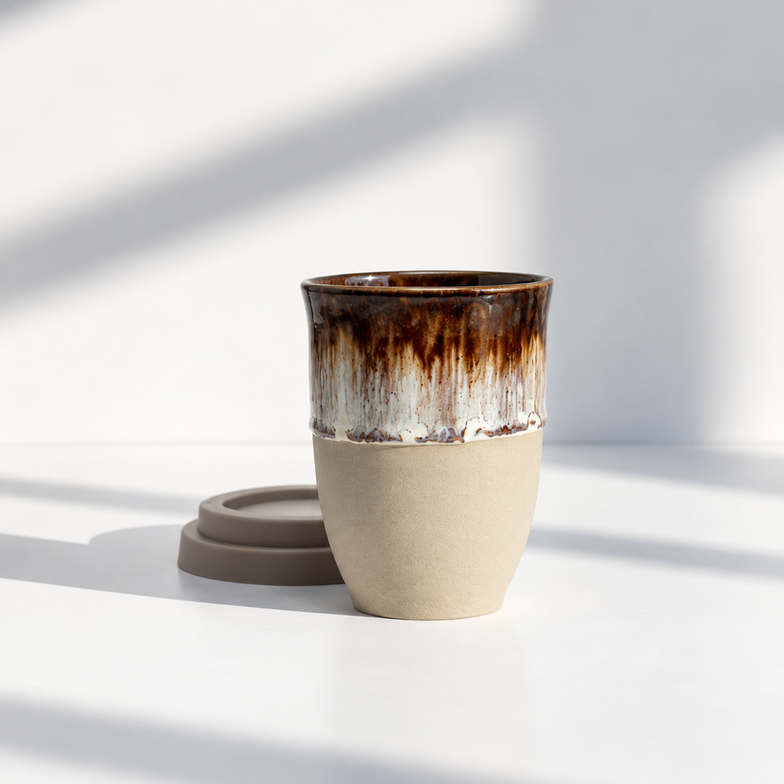 A ceramic travel mug with a textured beige lower half and a glossy brown dripping glaze upper half, shown with its matching brown lid on a white surface with soft window shadows.