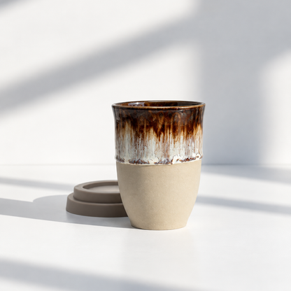 A ceramic travel mug with a textured beige lower half and a glossy brown dripping glaze upper half, shown with its matching brown lid on a white surface with soft window shadows.