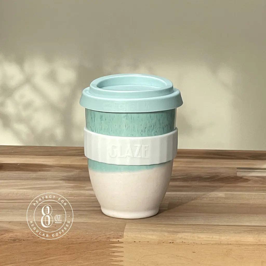 A reusable ceramic coffee cup with a light blue lid and band, a white middle band embossed with 'GLAZE', and a white lower body, sitting on a wooden tabletop against a beige wall.