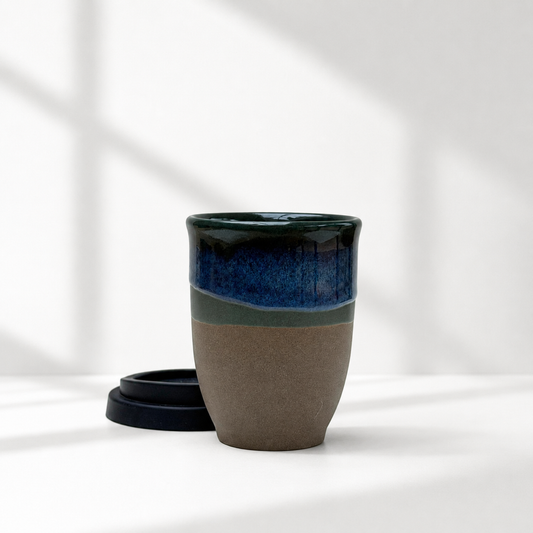A ceramic travel mug with a dark blue and green glazed top half and a matte brown bottom half, shown with its black lid on a white surface with soft window shadow overlays.