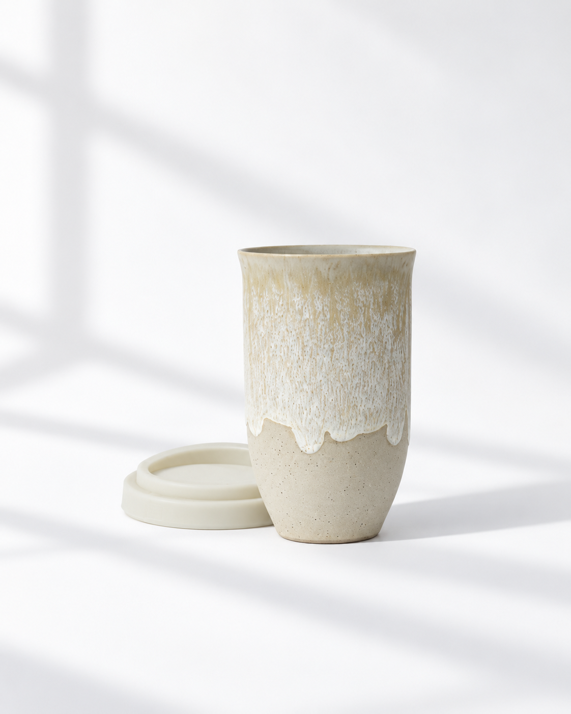 A ceramic travel mug with a matching lid, featuring a textured beige and white drip-glaze finish, set against a bright white background with soft window shadow patterns.