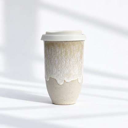 A ceramic travel tumbler with a white lid, featuring a textured cream and beige glaze with a dripping effect, set against a bright white background with soft diagonal shadows.