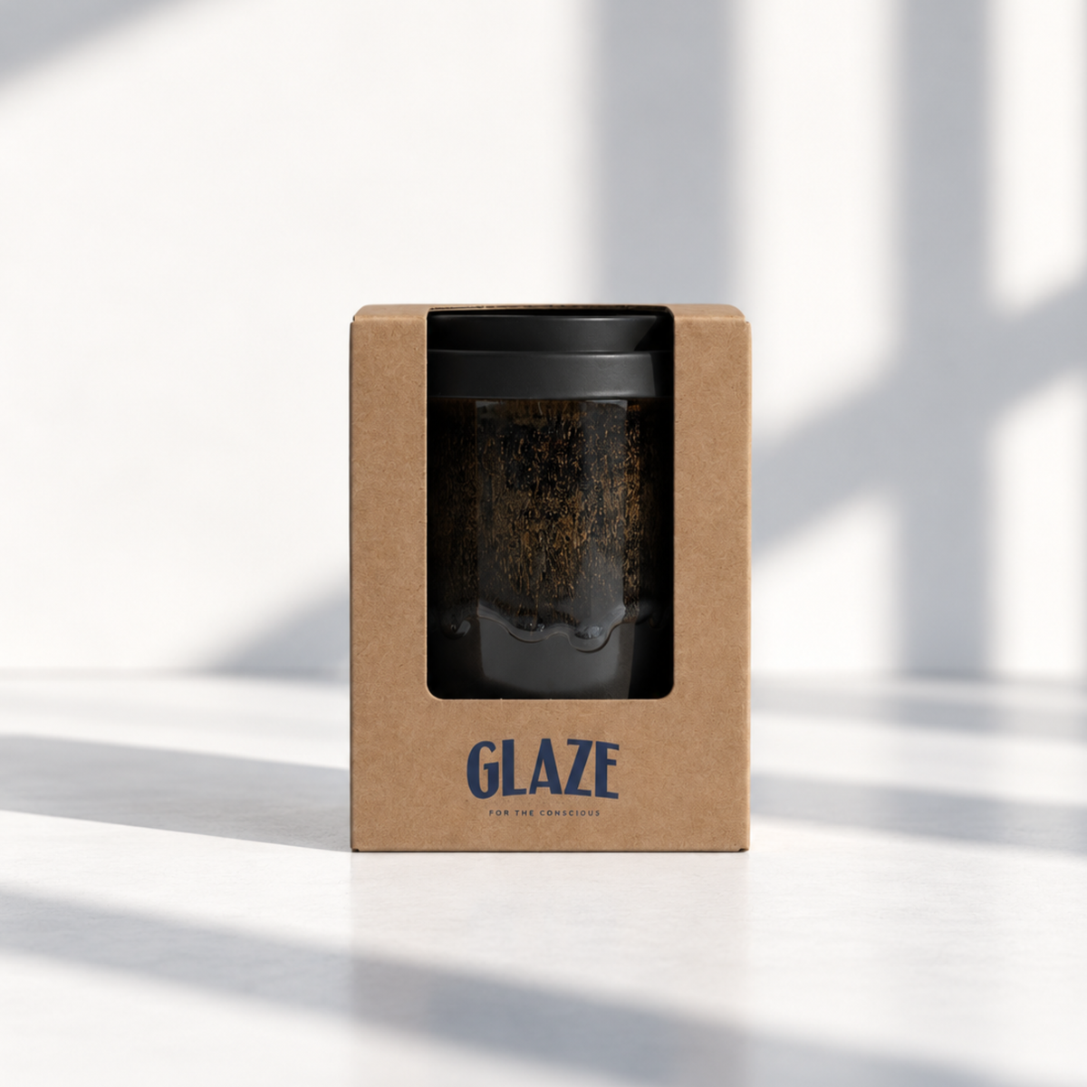 A reusable travel mug in its kraft paper packaging box, featuring a black window that displays the dark glass mug with gold speckles and a black lid. The box is branded with 'GLAZE' in blue text.