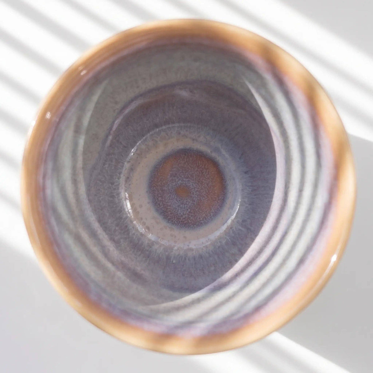 A top-down view of a ceramic cup or bowl with a textured, concentric glaze in shades of purple, gray, and warm brown, set against a white background with soft diagonal shadows.