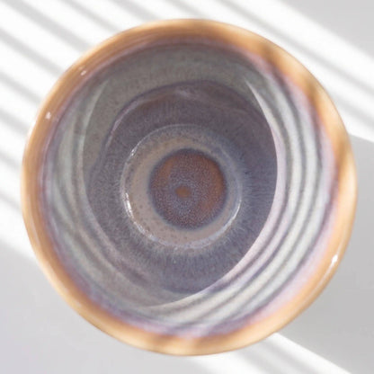 A top-down view of a ceramic cup or bowl with a textured, concentric glaze in shades of purple, gray, and warm brown, set against a white background with soft diagonal shadows.