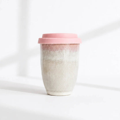 A reusable ceramic travel mug with a pink lid, featuring a textured, speckled glaze that transitions from a warm pink at the top to a light beige at the bottom, set against a bright white background with soft shadows.