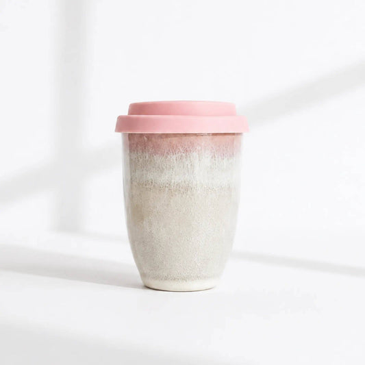 A reusable ceramic travel mug with a pink lid, featuring a textured, speckled glaze that transitions from a warm pink at the top to a light beige at the bottom, set against a bright white background with soft shadows.