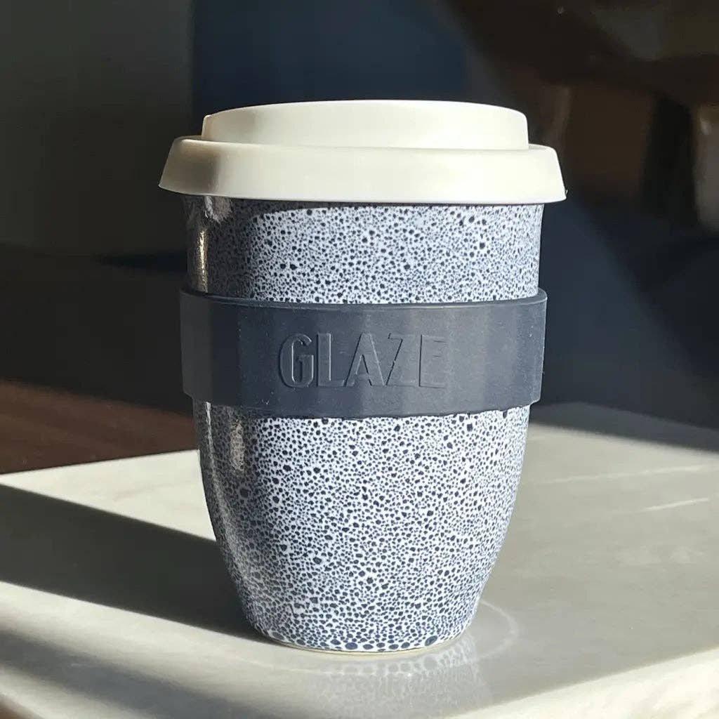 A reusable travel tumbler with a speckled light blue and white pattern, a dark gray silicone sleeve embossed with the brand name 'GLAZE', and a white lid, resting on a light-colored surface.