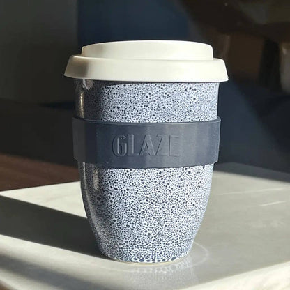 A reusable travel tumbler with a speckled light blue and white pattern, a dark gray silicone sleeve embossed with the brand name 'GLAZE', and a white lid, resting on a light-colored surface.