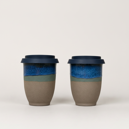 Two reusable ceramic travel mugs with dark blue lids, featuring a glossy blue upper band, a thin muted green stripe, and a matte taupe lower body, set against a plain light gray background.