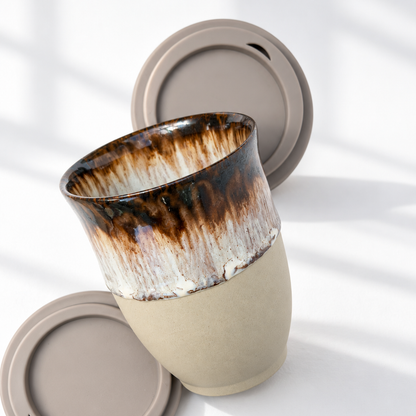 A ceramic reusable mug with a brown and cream glaze on the upper half and a matte beige lower half, shown with two matching beige lids on a white surface with soft window shadows.