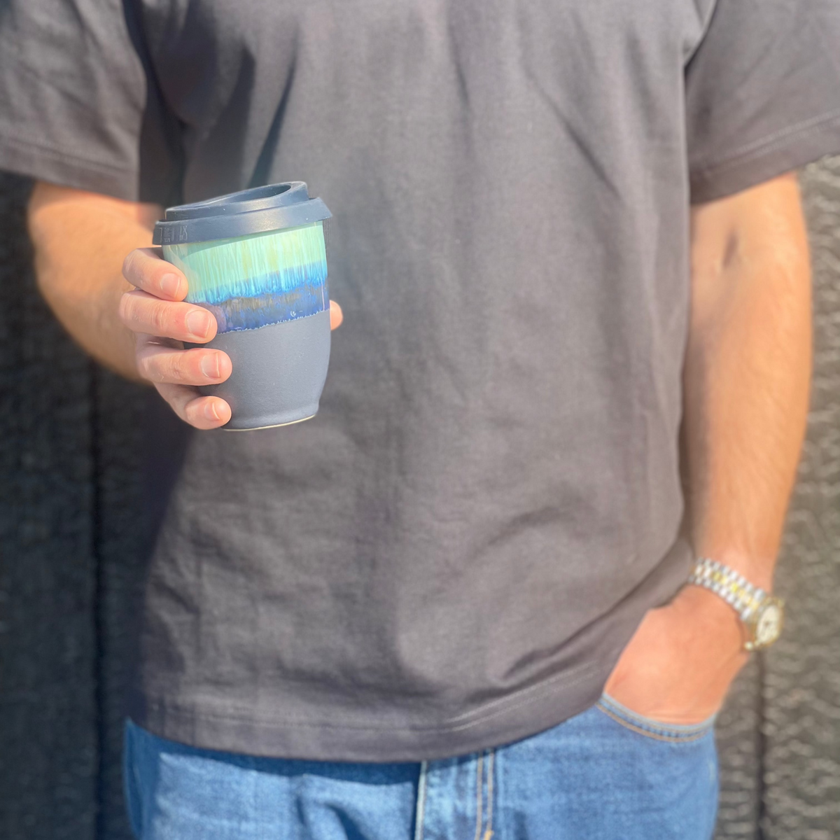 A person in a dark gray t-shirt holding a reusable coffee cup with a blue and green gradient band and a gray sleeve, set against a dark textured wall.