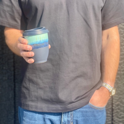 A person in a dark gray t-shirt holding a reusable coffee cup with a blue and green gradient band and a gray sleeve, set against a dark textured wall.