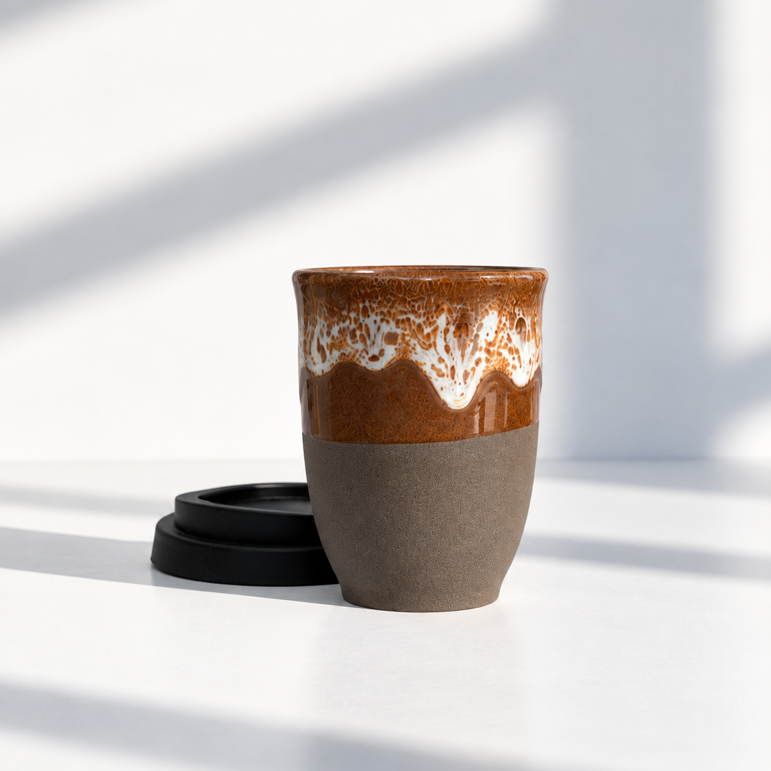 A ceramic travel mug with a glossy brown and white dripping glaze on the upper half and a matte taupe-brown lower half, shown with a black lid on a white surface in bright natural light.