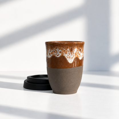 A ceramic travel mug with a glossy brown and white dripping glaze on the upper half and a matte taupe-brown lower half, shown with a black lid on a white surface in bright natural light.