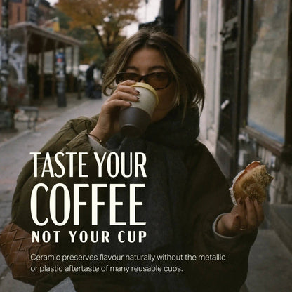 A woman on a city street drinks from a reusable coffee cup while holding a sandwich. Large overlaid text reads 'TASTE YOUR COFFEE NOT YOUR CUP' with smaller descriptive copy below, suggesting a promotional image for a coffee cup product.