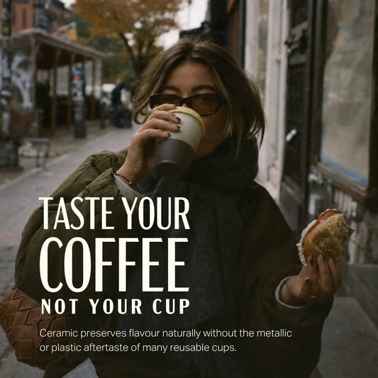A woman on a city street drinks from a reusable coffee cup while holding a sandwich. Large overlaid text reads 'TASTE YOUR COFFEE NOT YOUR CUP' with smaller descriptive copy below, suggesting a promotional image for a coffee cup product.