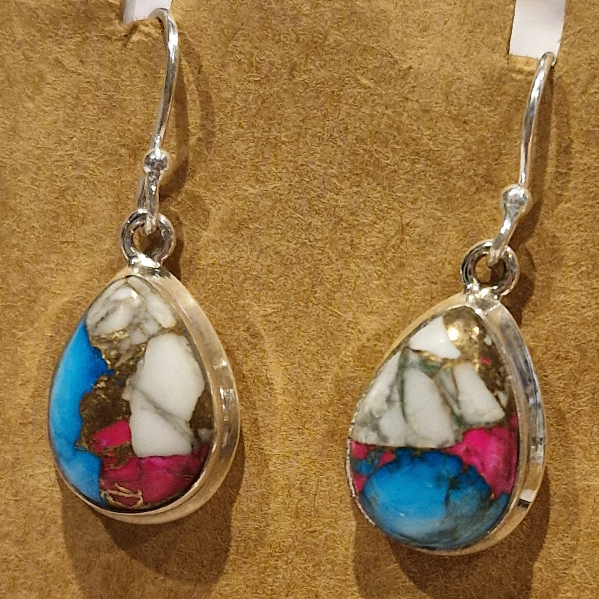 A pair of teardrop-shaped silver dangle earrings featuring a multicolored turquoise-like stone inlay with blue, white, pink, and brown marbling, displayed on a textured tan background.