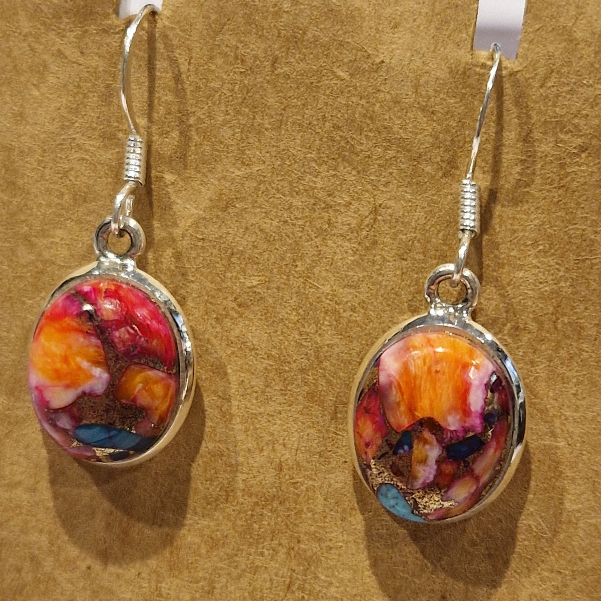 A pair of oval dangle earrings with colorful abstract marbled resin centers in red, orange, pink, yellow, and blue, set in silver-tone metal with silver fishhook wires, displayed on a textured tan background.