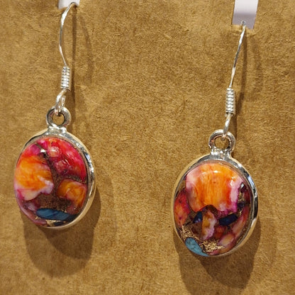 A pair of oval dangle earrings with colorful abstract marbled resin centers in red, orange, pink, yellow, and blue, set in silver-tone metal with silver fishhook wires, displayed on a textured tan background.